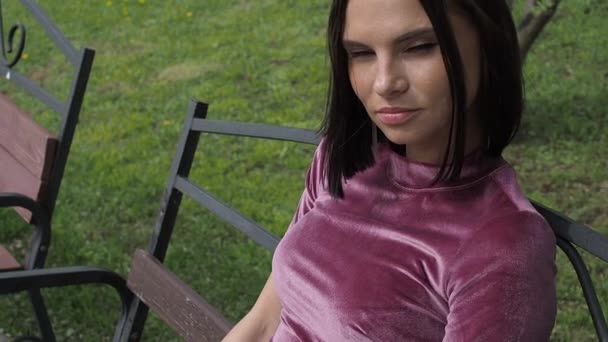 エレガントな若いブルネットリラックス上のベンチで公園 - 映像、動画