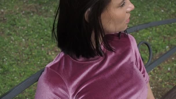 Κομψή νεαρή μελαχρινή χαλαρώνοντας στον πάγκο στο πάρκο - Πλάνα, βίντεο