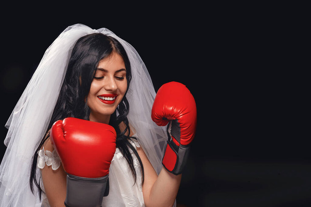 portrait attractive brunette with red lipstick, in wedding dress, veil and Boxing gloves. young girl in role bride on black background, clasping her hands on her chest, protects herself and looks away - Photo, Image