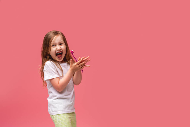 a happy little girl enjoying brushing her teeth, laughing with healthy white teeth and showing a gesture of approval, class on a pink background. Oral hygiene. Daily brushing of teeth, 2 times a day. - 写真・画像