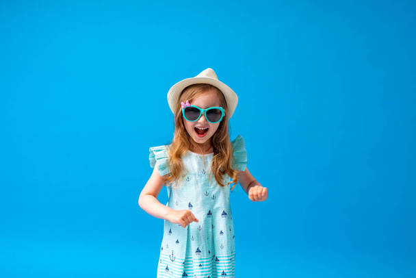cute little girl 4 years old, in a dress, hat and sunglasses, posing on a blue background. The child will be happy to start the summer holidays and go on a trip - Foto, Imagen