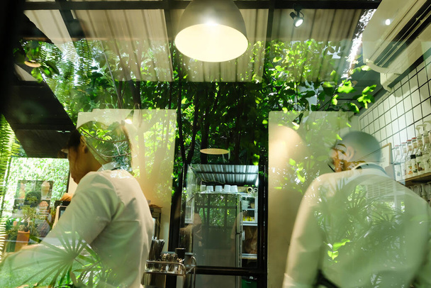 Vista a través de la ventana de la cafetería verde, jóvenes baristas femeninos y masculinos en uniformes blancos trabajando en una cafetería moderna
. - Foto, imagen