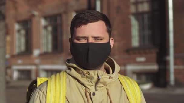 Caucasian male delivery man in a black mask and a jacket with a yellow backpack turns to the camera, takes off his mask and takes a deep breath. - Video