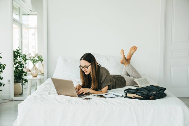 Studentessa positiva si trova a letto e utilizza un computer portatile. Sta messaggiando o navigando online su Internet. - Foto, immagini