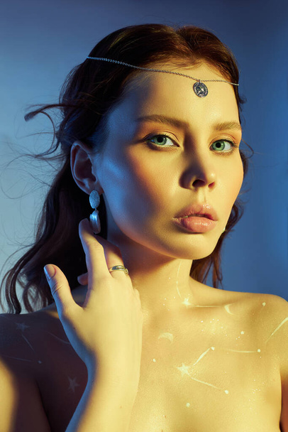 Beauty portrait of a woman with beautiful makeup, earrings and a necklace on the girl. Drawing Cosmos constellation of the zodiac on a woman chest - Photo, Image