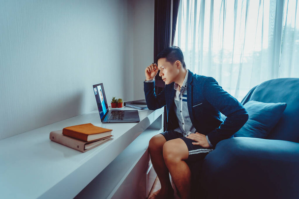Jóvenes empresarios asiáticos usan traje formal con pantalones cortos que tienen una reunión de videoconferencia con su socio de negocios en el sofá en casa en la sala de estar durante el período de cuarentena
. - Foto, Imagen