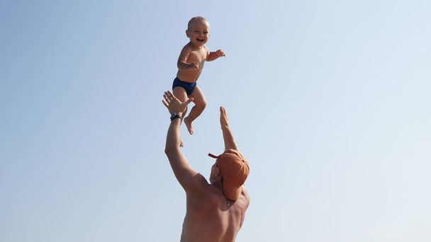 Ένα μικρό χαρούμενο παιδί και ο αθλητικός του πατέρας στην παραλία. Ο μπαμπάς ξέρασε γελαστό αγόρι ψηλά στον αέρα. - Φωτογραφία, εικόνα