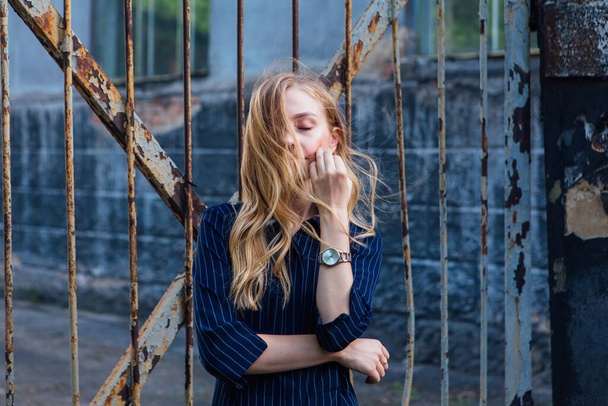 Close up portrait of a fashion look's woman with long blond hair standing near the old rusty gate. Young woman modern portrait. - Photo, Image