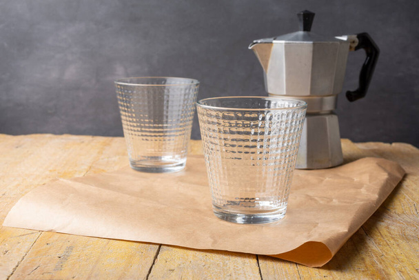 Nahaufnahme zweier leerer Gläser mit grauer Kaffeekanne auf braunem Papier auf Holztisch, grauer Hintergrund, waagerecht, mit Kopierraum - Foto, Bild