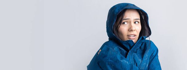 Egy kék esőkabátba öltözött mosolygós lány portréja, amint csuklyával pózol a szürke háttéren egy stúdióban.. - Fotó, kép