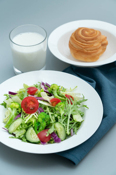 Ένα πιάτο με υγιεινή σαλάτα και πρωινό ψωμί.Ένα πιάτο με νόστιμη και υγιεινή σαλάτα λαχανικών σε ένα γκρι τσιμεντένιο πάτωμα. - Φωτογραφία, εικόνα