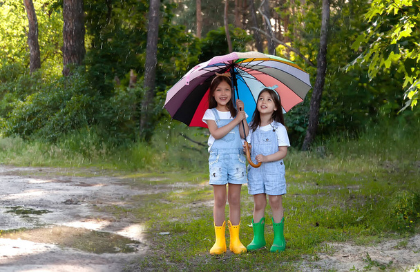 Girls sister friends in denim overalls and colorful rubber boots smile at the camera. Summer rain and a rainbow umbrella Laugh and enjoy life. Siblings family. Positive and full of life - Photo, Image