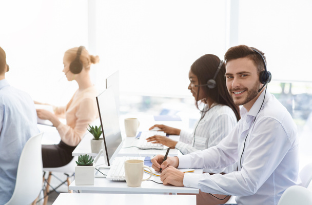 Aantrekkelijke call center operator glimlachend naar de camera en zijn collega 's praten met klanten op kantoor, kopieer ruimte - Foto, afbeelding