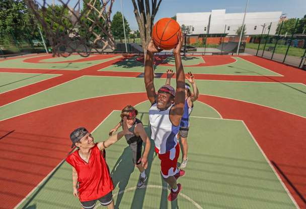 Jugador de baloncesto negro saltando para marcar gol y su equipo en la cancha al aire libre
 - Foto, Imagen