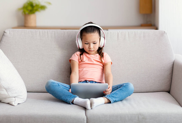 Tyttö käyttää digitaalinen tabletti ja kuulokkeet katsomassa piirrettyjä istuu sisätiloissa - Valokuva, kuva
