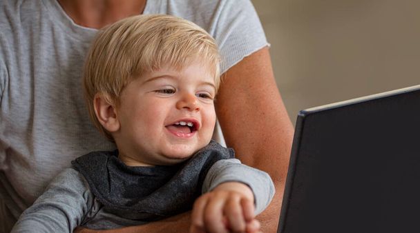 Pequeño niño mirando emocionado por algo en un monitor de computadora portátil en el regazo de la madre
. - Foto, imagen