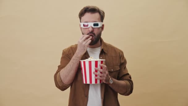 An attractive focused young unshaven man with 3d eyeglasses is eating popcorn while watching something excited isolated over beige wall background in studio - Felvétel, videó