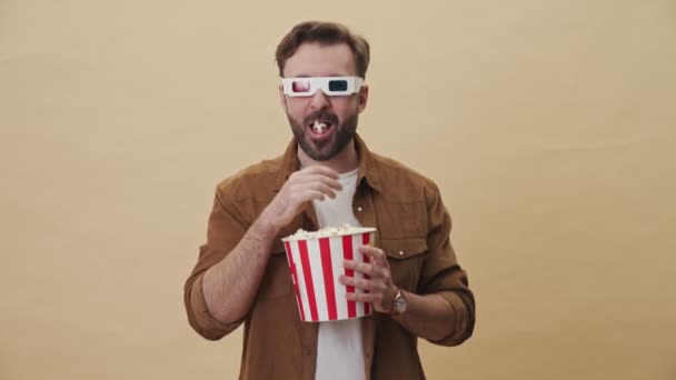 A happy young unshaven man with 3d eyeglasses is eating popcorn while watching something excited and laughing isolated over beige wall background in studio - Séquence, vidéo