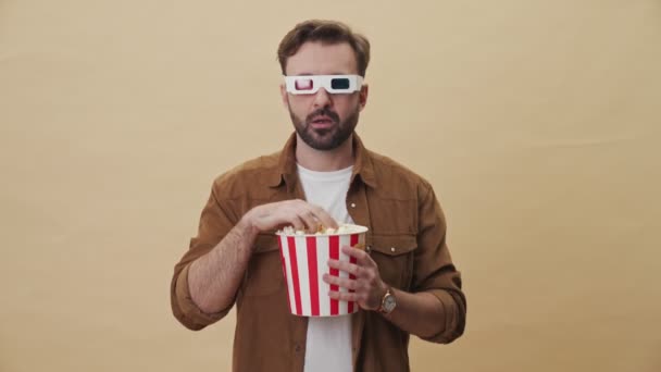 A frightened young unshaven man with 3d eyeglasses is eating popcorn while watching something scary isolated over beige wall background in studio - Felvétel, videó