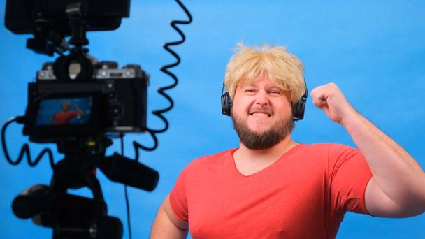 hátborzongató kövér férfi parókában és rózsaszín pólóban csinál egy videó blog a kék háttér. - Fotó, kép