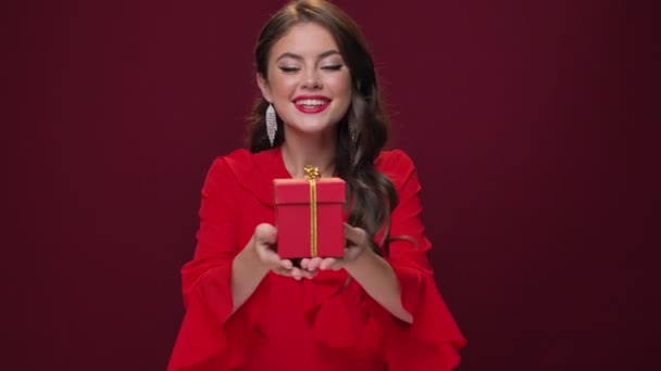 A friendly young woman wearing red dress is giving a present to the camera holding it in her hands isolated over burgundy background - Video