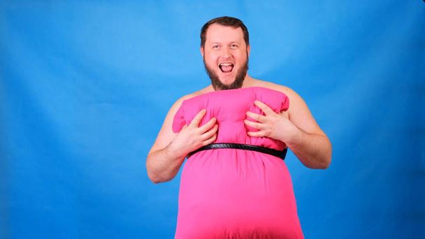 Funny bearded guy in a pink dress made of pillows touches his chest on a blue background. Crazy quarantine. Funny house cleaning. Fashion 2020. Put on a pillow. Challenge 2020 due to house isolation - Foto, Imagen