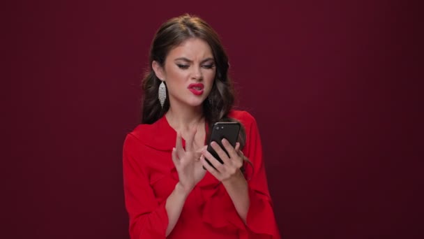 An emotional pretty nice young woman wearing red dress is choosing something in her smartphone while holding it in her hands isolated over burgundy background - Felvétel, videó