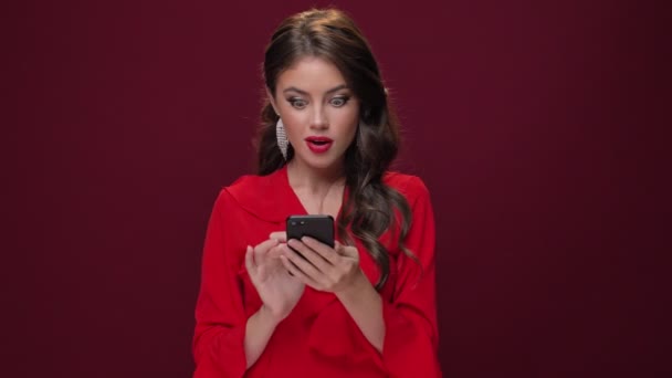 A happy cheerful young woman wearing a red dress is getting brilliant news and doing a winner gesture isolated over burgundy background - Felvétel, videó