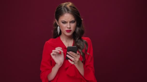 An attractive young woman wearing a red dress is watching bored and bad content on her smartphone isolated over burgundy background - Filmati, video