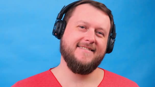 Bearded funny cute dikke man in een roze T-shirt luistert naar muziek en danst op een blauwe achtergrond, close-up, kopieer ruimte. - Video