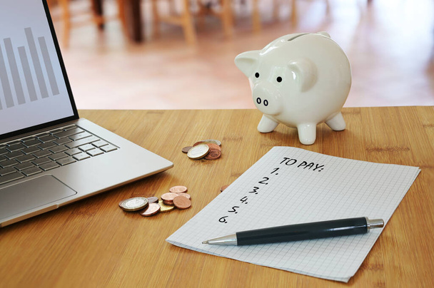 Calculating household costs, laptop, coins, a piggy bank and a list what to pay on a table in the living room, economy finance concept to save money, selected focus, narrow depth of field - Foto, afbeelding