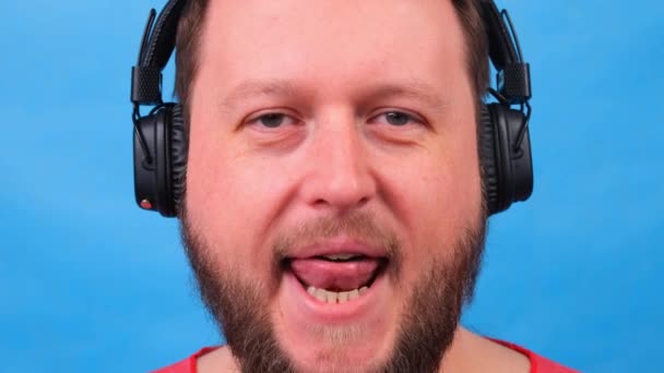 Bearded funny cute fat man in a pink T-shirt listens to music and dances and shows his tongue on a blue background, close-up, copy space. - Footage, Video