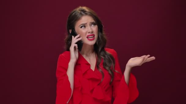 Kırmızı elbiseli kızgın genç bir kadın, bordo arka planda izole bir şekilde akıllı telefonuyla konuşurken biriyle tartışıyor. - Video, Çekim