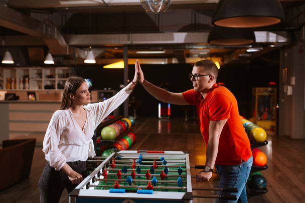Piękna biała dziewczyna i mężczyzna w okularach grają w futbol stołowy w pokoju zabaw i dają pięć. Miłość i rekreacja - Zdjęcie, obraz