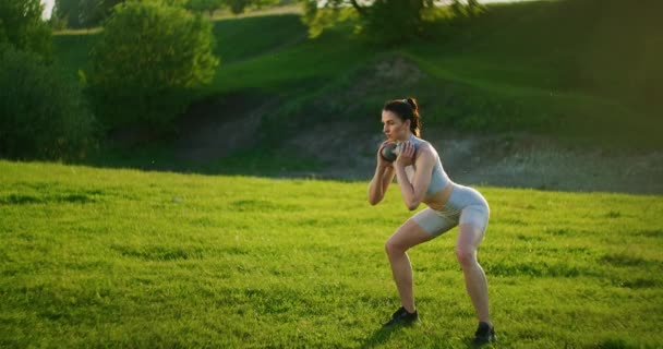 Kobieta przykuca z hantlami w parku o zachodzie słońca i rzuca się na bok. Motywacja i treningi sportowe. Samotna kobieta ćwiczy o zachodzie słońca w lecie - Materiał filmowy, wideo