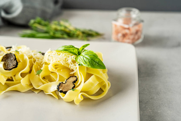 Fettuccine pasta with black truffle and grated Parmesan cheese is served on a beige rectangular plate, decorated with fresh basil leaves. Light gray concrete background, close-up. - Fotoğraf, Görsel