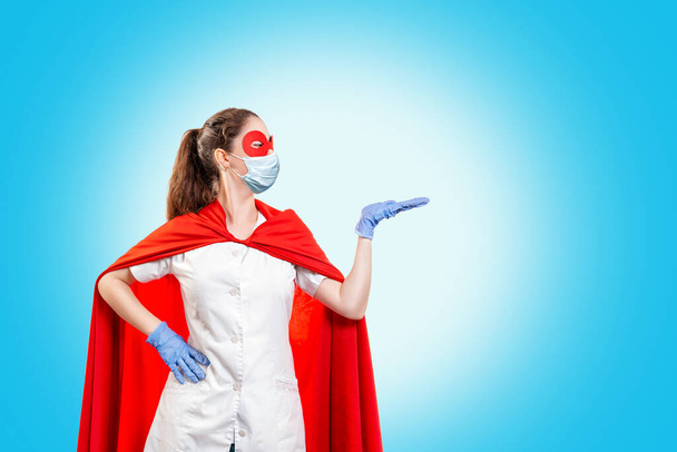 Ein Arzt mit medizinischer Maske, Handschuhen und rotem Cape zeigt zur Seite. Blauer Hintergrund. Kopierraum. Das Konzept der Macht eines Superhelden für Medizin und Gesundheitswesen. - Foto, Bild
