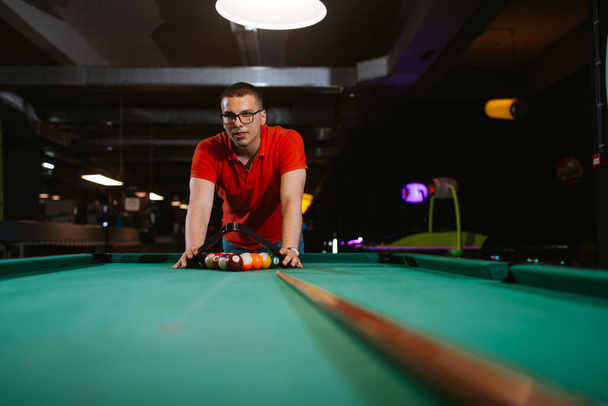 Un jeune homme caucasien en t-shirt rouge place des balles sur une table de billard. Temps de plaisir  - Photo, image