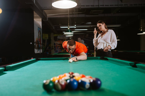 A young caucasian man in a red T-shirt is playing billiards while a girl is standing next to him. Fun time - Foto, immagini