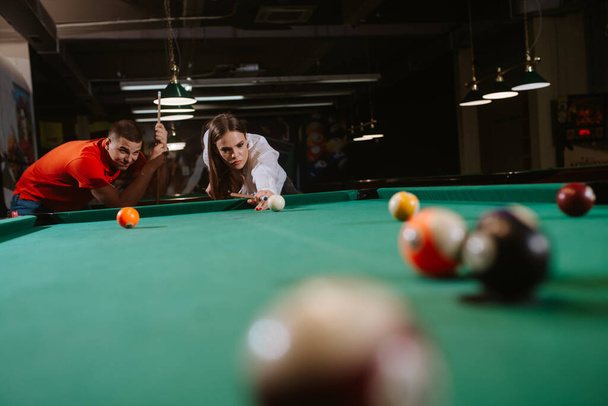 Μια νεαρή λευκή γυναίκα και ένας λευκός παίζουν μπιλιάρδο. Μια όμορφη γυναίκα σημαδεύει μια μπάλα, ενώ ένας άντρας με κόκκινο μπλουζάκι στέκεται δίπλα της. Ώρα για διασκέδαση - Φωτογραφία, εικόνα