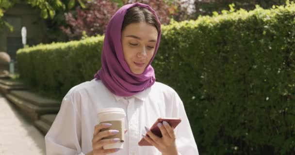 Caminhando bela jovem bebe café e olhando para o smartphone, vestindo lenço de cabeça tradicional. Mulher atraente no hijab
 - Filmagem, Vídeo