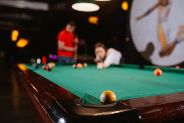 A young caucasian woman and a caucasian man play billiards. A beautiful woman is aiming a ball, while a man in a red T-shirt is standing next to her. Fun time - Photo, Image