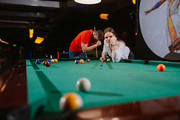 Egy fiatal kaukázusi nő és egy fehér férfi biliárdozik. Egy gyönyörű nő egy labdát céloz, miközben egy piros pólós férfi áll mellette. Szórakozás. - Fotó, kép