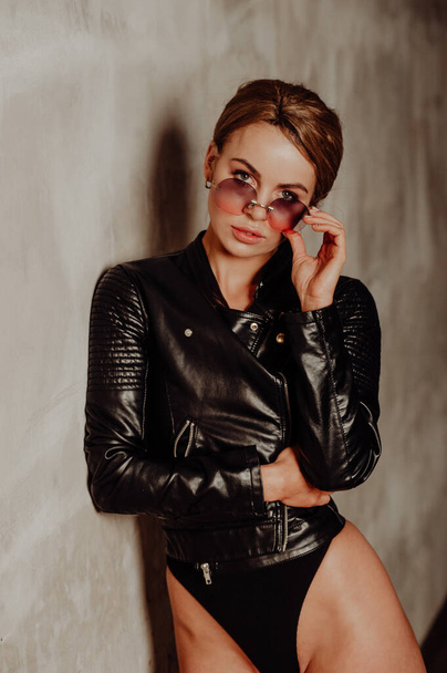 Rock concept. Portrait of charming blond woman in black bodysuit and leather jacket. Pink sunglasses.  - Foto, imagen