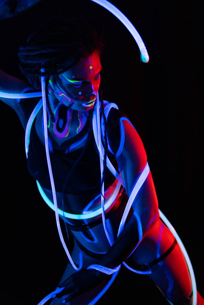 Portrét Warror Girl s žhnoucími trubicemi v Neon UF Light. Modelka s dredy a fluorescenčním kreativním psychedelickým make-upem, design ženského diskotékového modelu v UV, barevné abstraktní - Fotografie, Obrázek