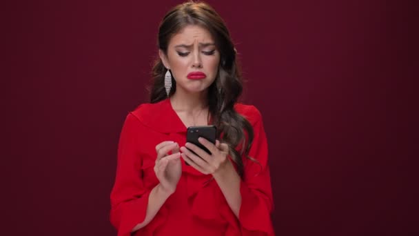 An emotional disappointed young woman wearing a red dress is becoming upset while watching something on her smartphone isolated over burgundy background - Video
