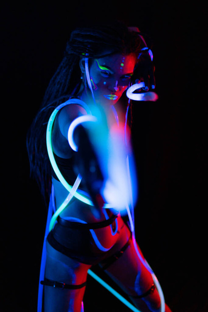 ネオンUFライトで輝くチューブを持つ戦争少女の肖像画。ドレッドロックと蛍光クリエイティブサイケデリックメイクアップ、 UVの女性ディスコダンサーモデルのアートデザイン、カラフルな抽象 - 写真・画像