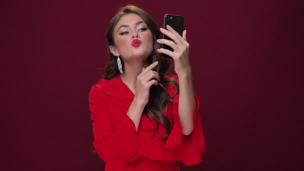 Kırmızı elbiseli güzel, memnun bir genç kadın kendine bakıyor. Burgonya arka planında izole edilmiş akıllı telefonundaki kamerayı kullanıyor. - Video, Çekim