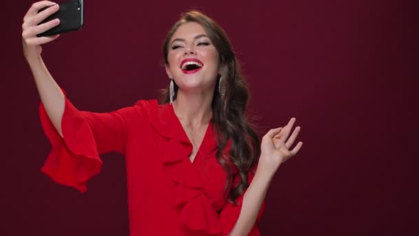 A fashionable attractive young woman wearing a red dress is taking selfie-photos using her smartphone isolated over burgundy background - Footage, Video
