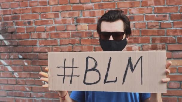 Кавказький чоловік у масці стоїть навпроти червоної стіни з картонним плакатом у руках з написом хештег - BLM. Єдиний протест. - Кадри, відео